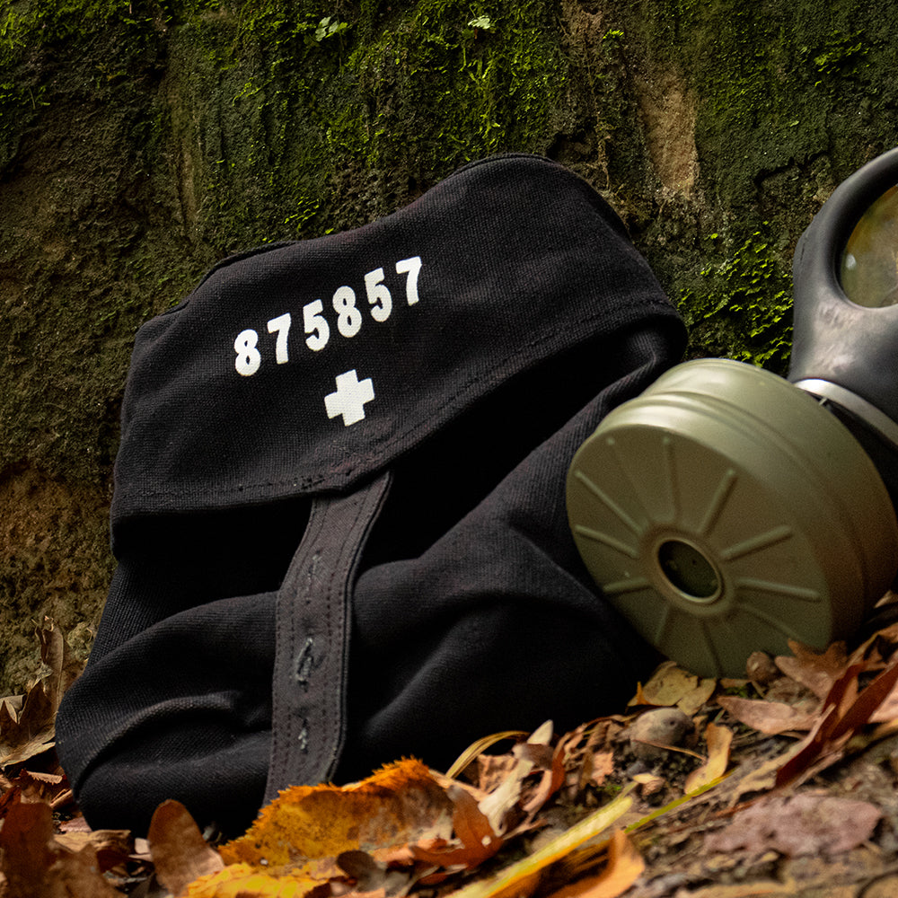 An empty Swiss Gas Mask Bag laying against a mossy rock on fallen leaves with a gas mask next to it.