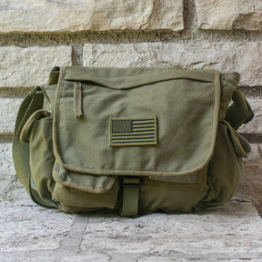 Olive Drab Retro Messenger Bag with U.S. Flag patch sat in front of a brick wall.