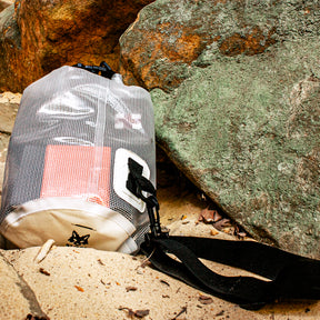 10 Liter Clear Dry Bag lying on a sandy bank surrounded by large rocks.