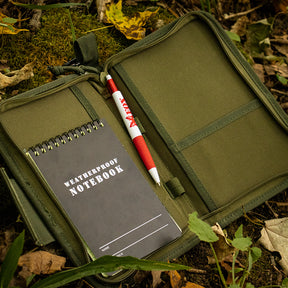 An open Field Notebook/Organizer Case laying on a mossy and leafy forest floor with a weatherproof notebook and pen inside.