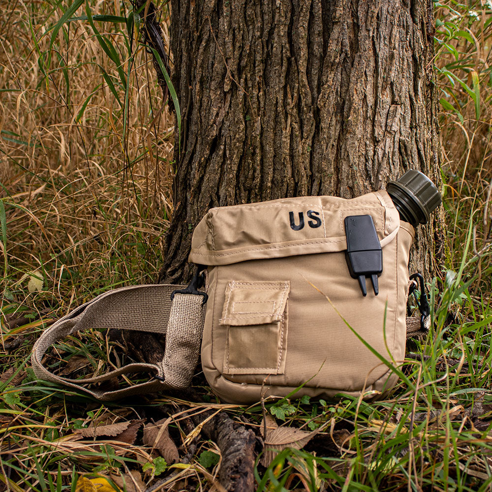 GI 2 Quart Canteen Cover with a canteen inside leans against a tree within a grassy field.
